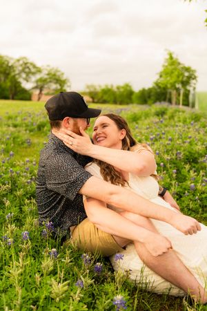 Happy couple sitting together in a field of bluebonnets, sharing a romantic moment on a cloudy day. irving engagement session