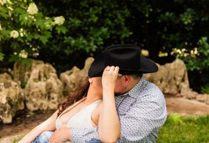 Couple sharing a romantic kiss hidden under a black cowboy hat outdoors near rocks and greenery. Dallas Arboretum Engagement Session