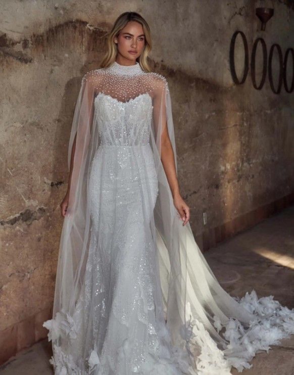 Elegant white lace wedding gown with sheer cape overlay and sweetheart neckline against textured wall.