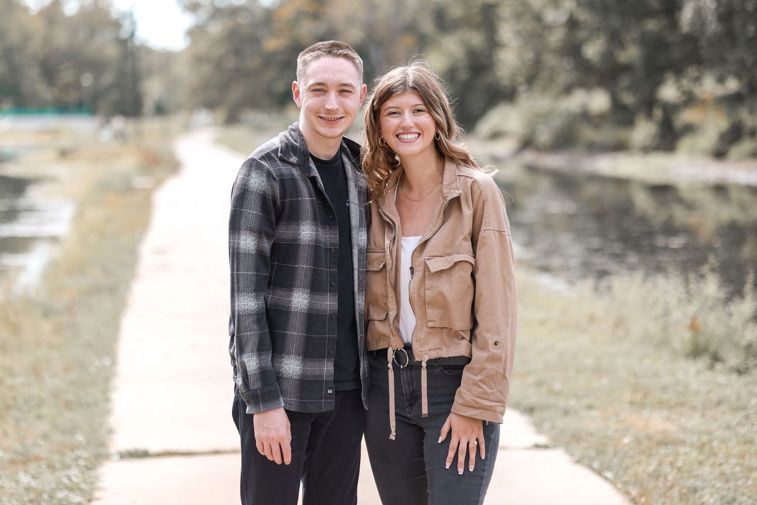 Couple poses together on a path near a serene pond wearing plaid and tan jackets.