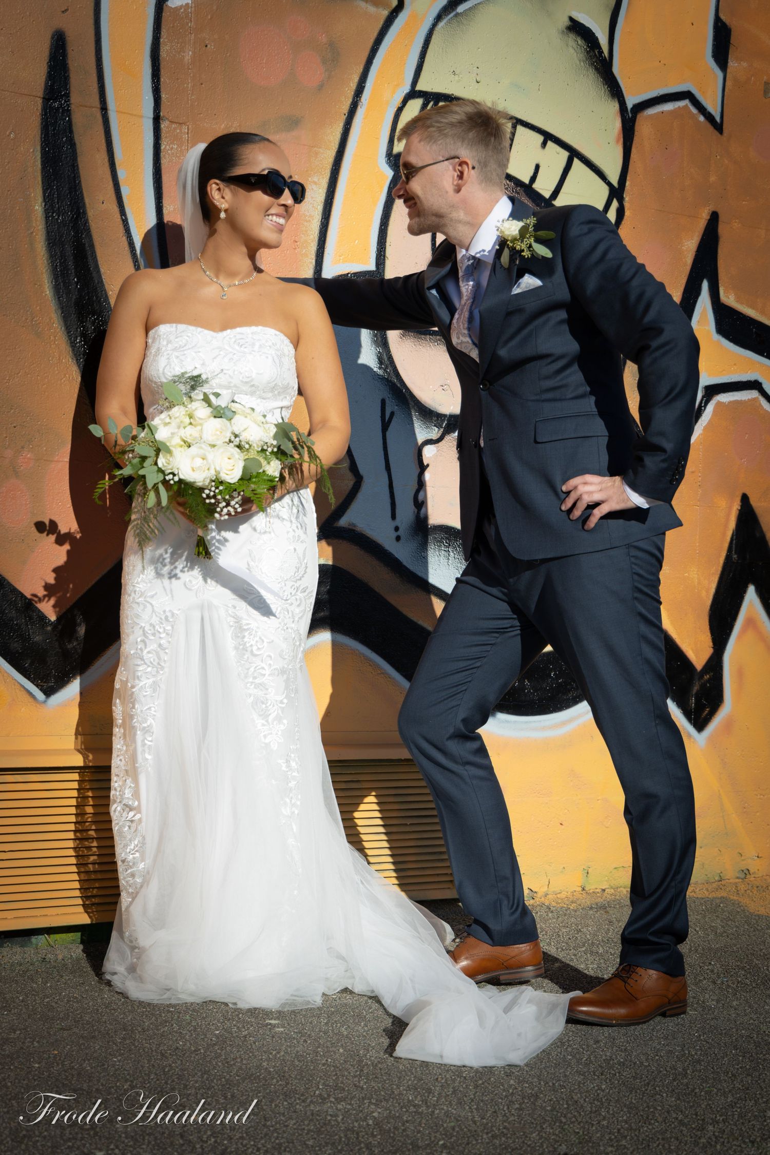 Laughing bride in sunglasses and groom posing playfully in front of colorful graffiti wall.