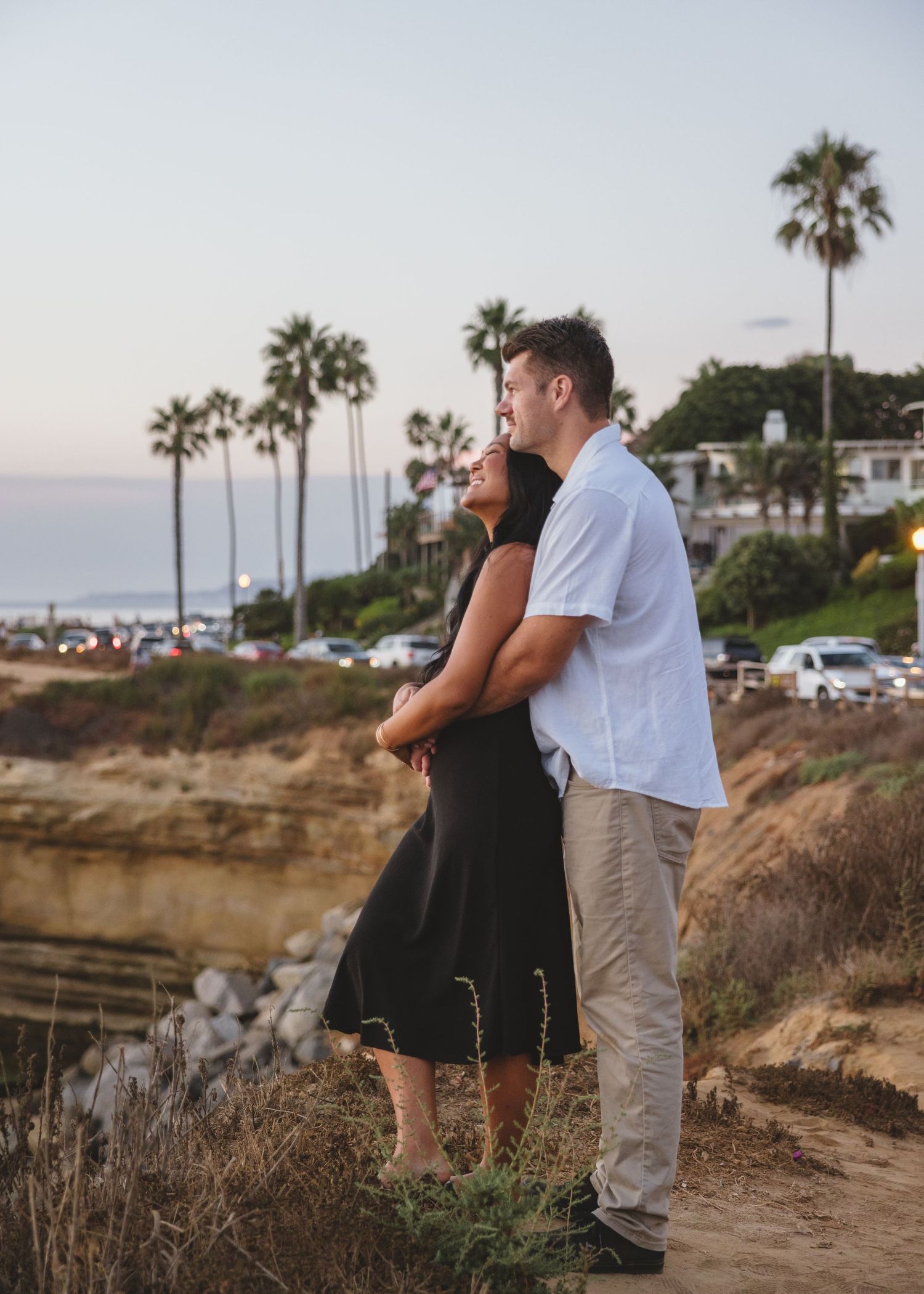 Couple embracing on coastal cliff path with palm trees and ocean views.