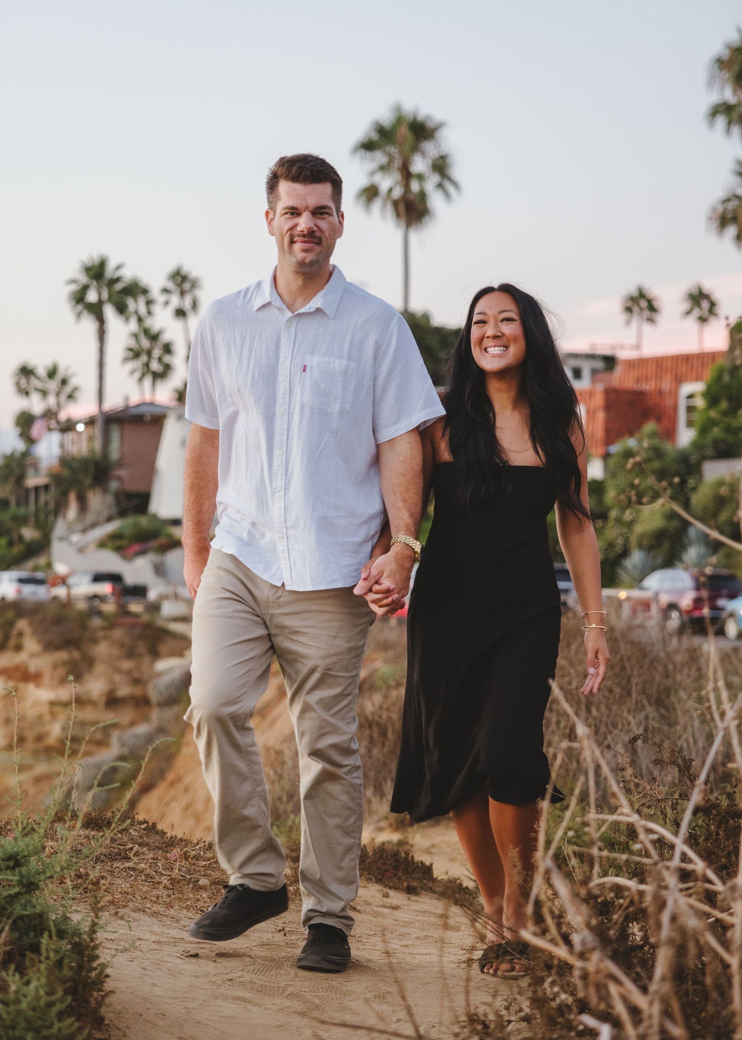 Couple walking hand in hand on coastal path with palm trees in background.
