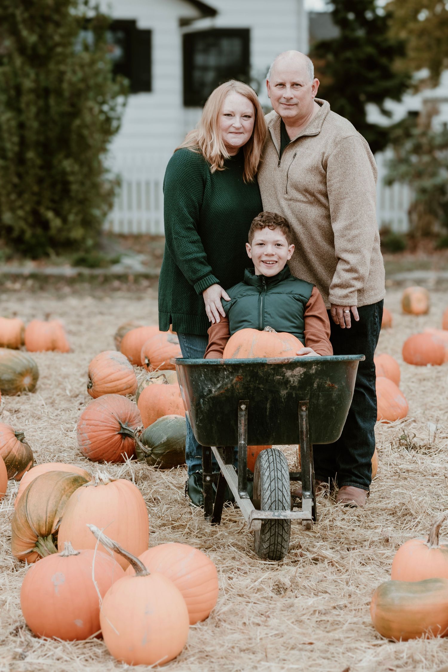 Family posing with wheelbarrow in pumpkin patch during fall season.