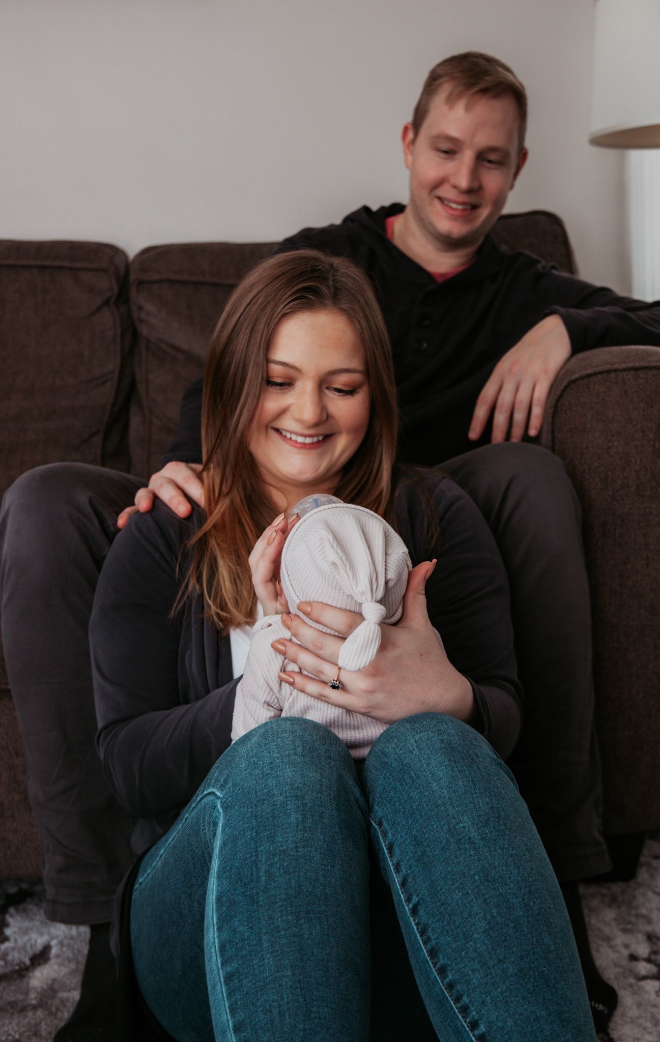 A couple relaxes on a brown couch while holding a newborn baby wrapped in white.