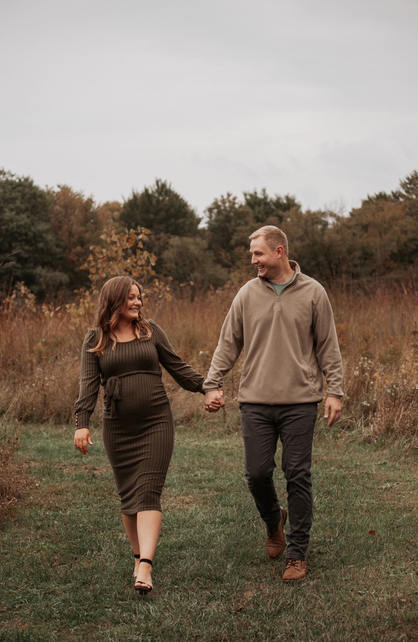 Couple walks hand in hand through autumn field during maternity photoshoot.