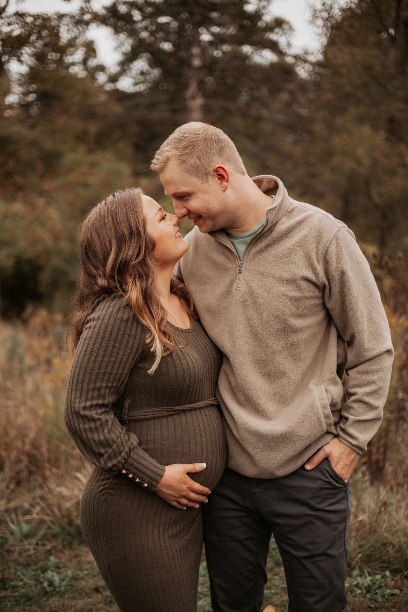 A tender moment between an expecting couple in a fall field wearing casual sweaters.