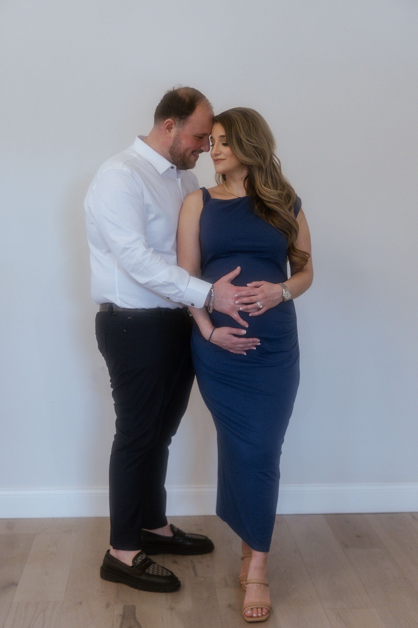 Couple in formal attire embraces lovingly against plain white wall.