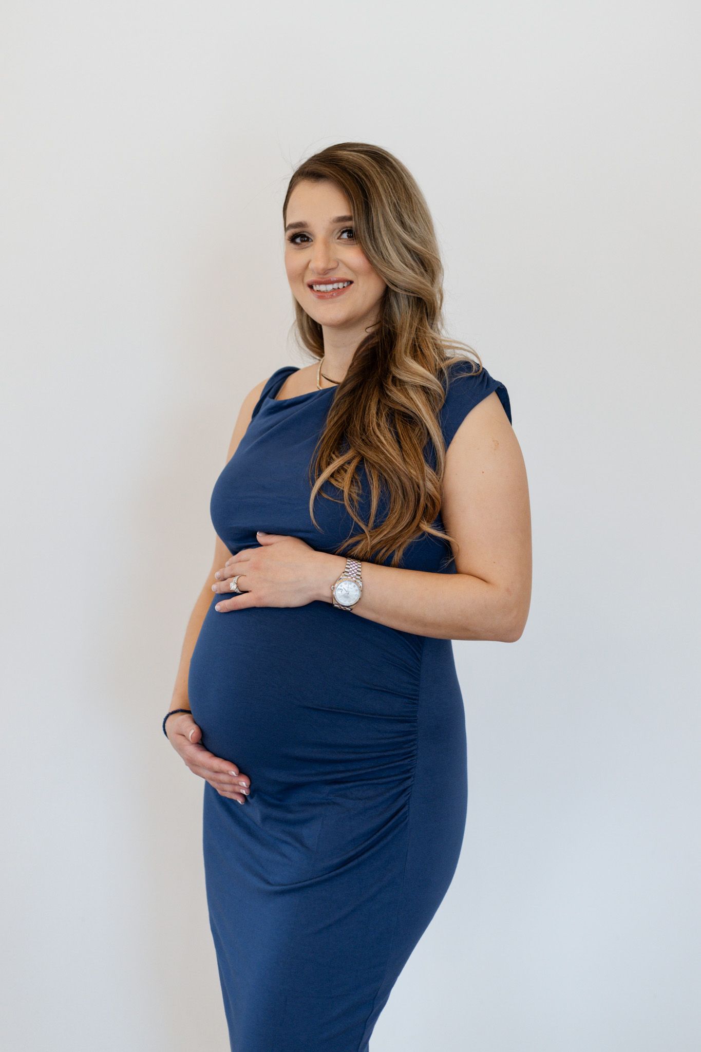 Expectant mother in navy blue dress poses for maternity photos against white backdrop.