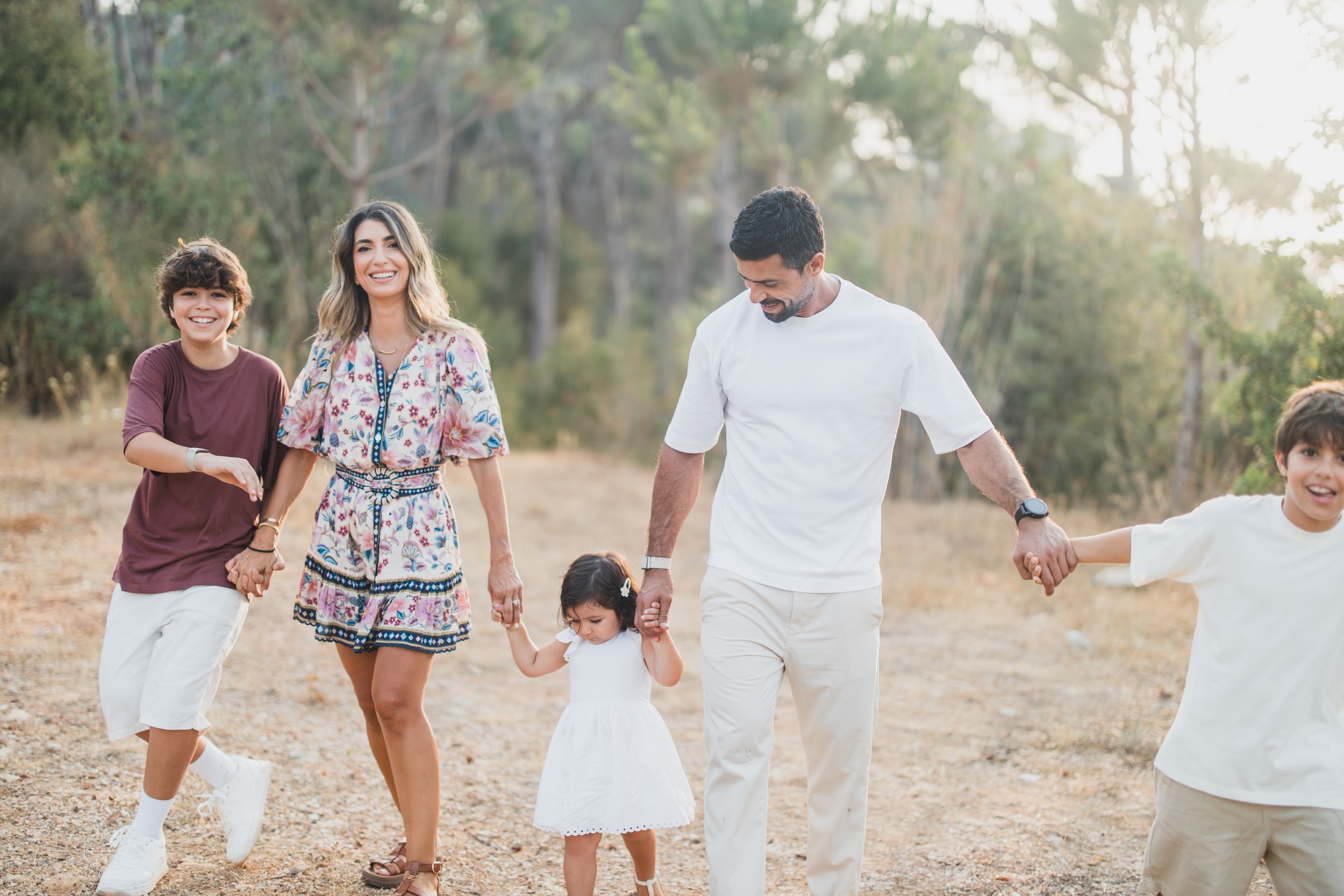 Family wearing coordinated white and burgundy outfits walking together through a sunlit forest setting.