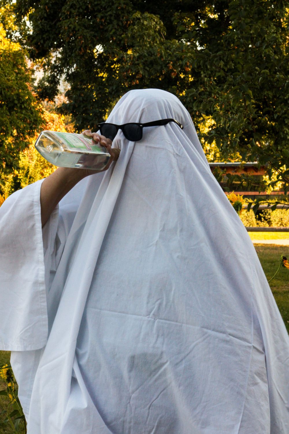 Figure in white ghost costume with sunglasses drinks from bottle in outdoor garden setting.