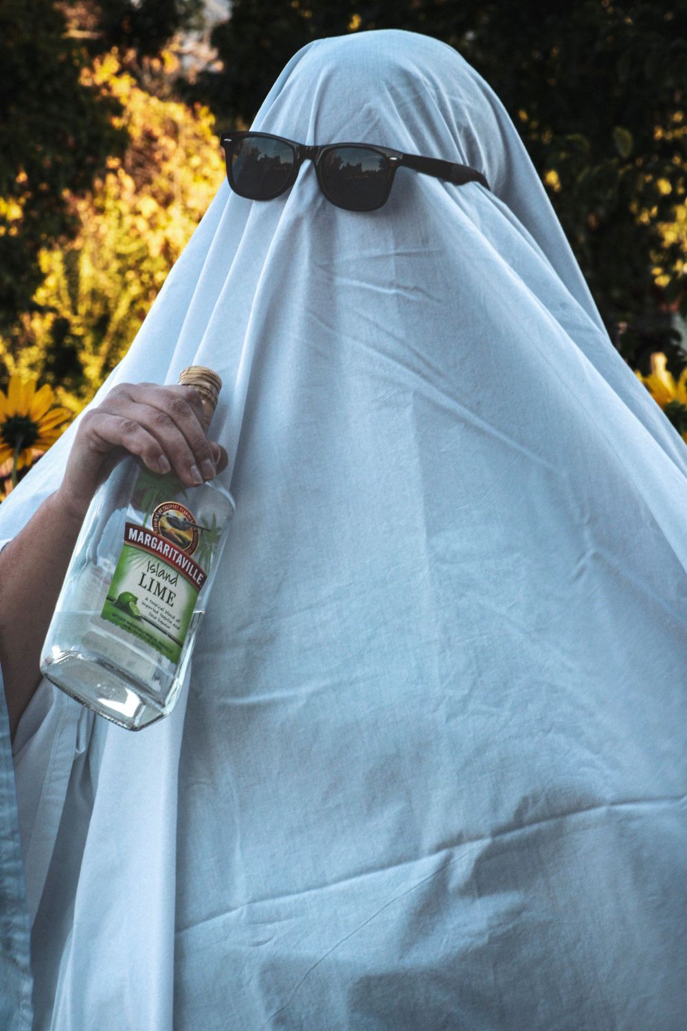 Ghost-like figure in sunglasses holding clear bottle against autumn foliage background.