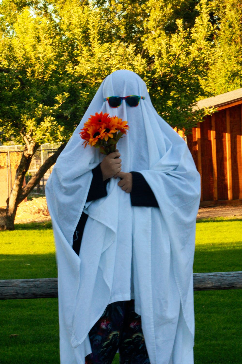 Ghost figure wearing sunglasses holds orange flowers in backyard garden setting.