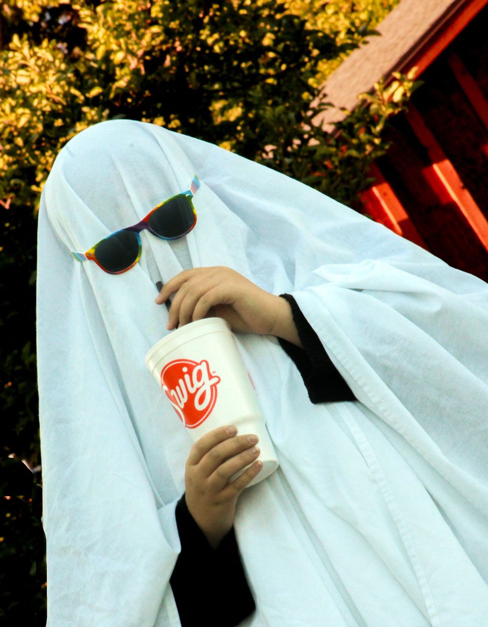 Playful ghost figure in sunglasses holds Sonic cup against autumn background.