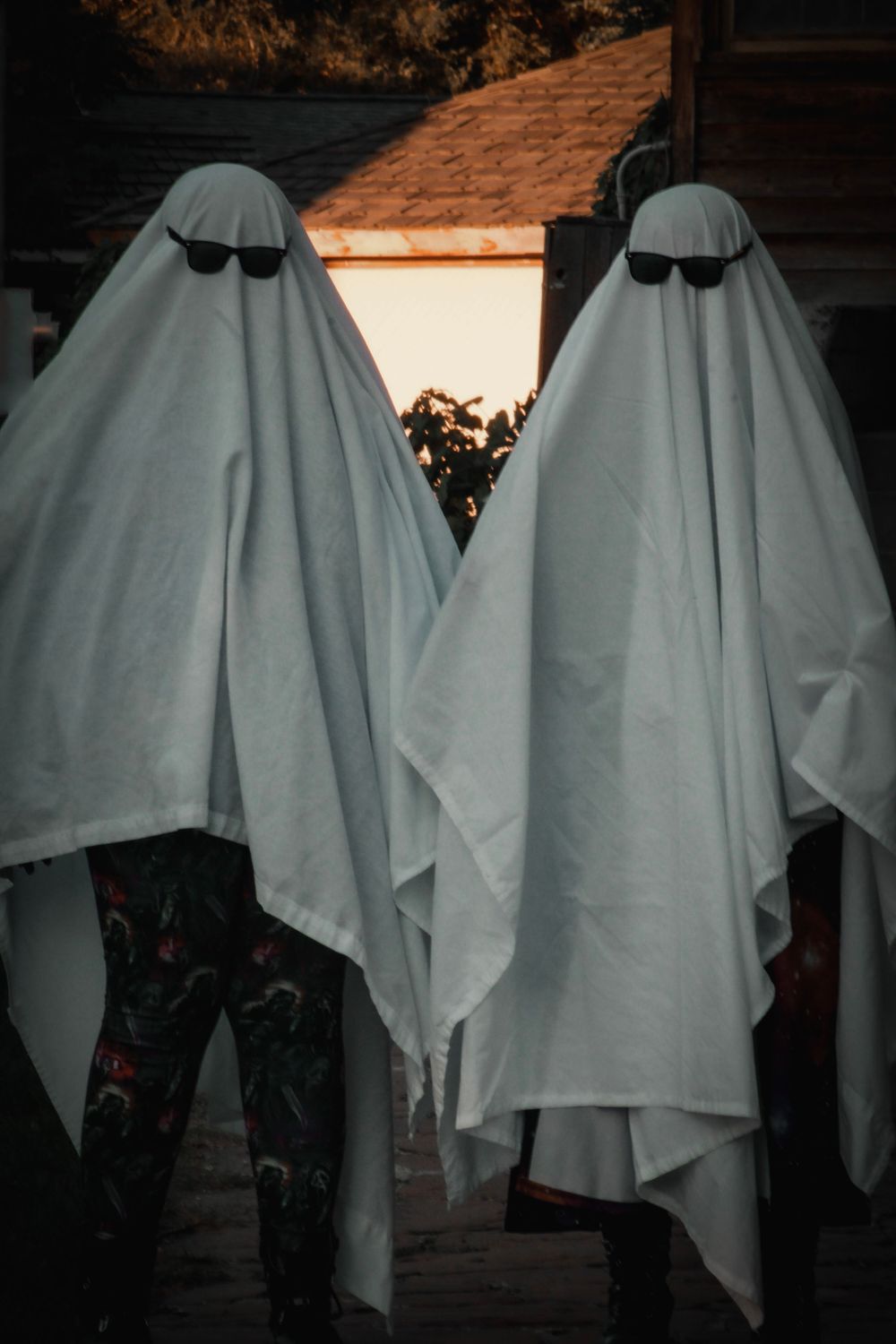 Two ghost costumes with sunglasses stand side by side against wooden building.