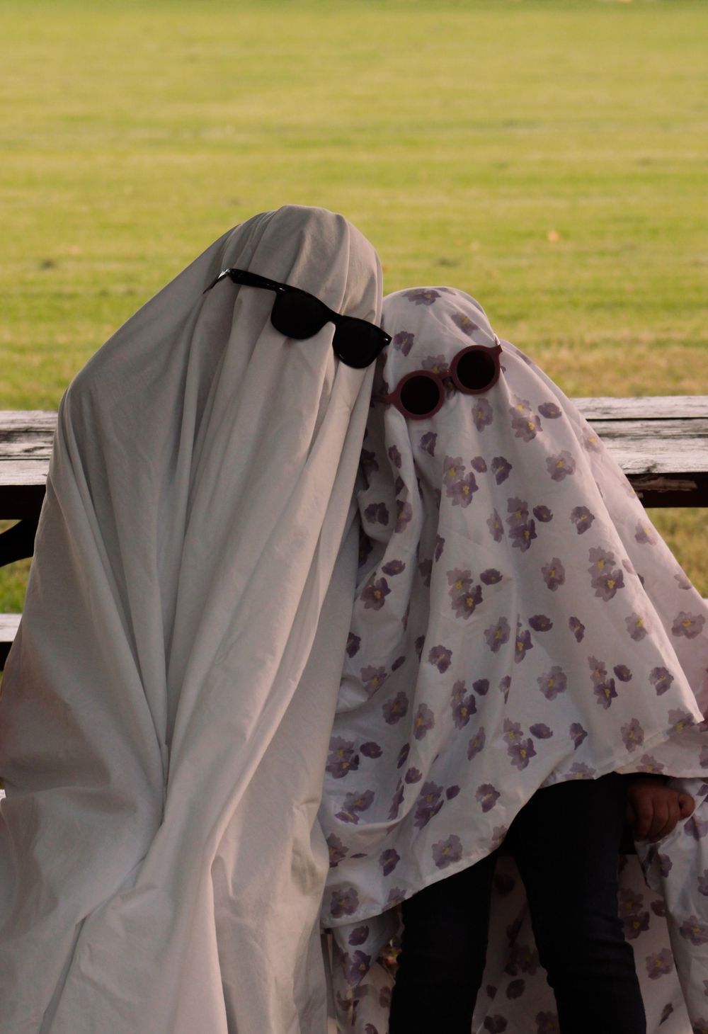 Two ghost figures in sunglasses sit together on wooden bench in field.