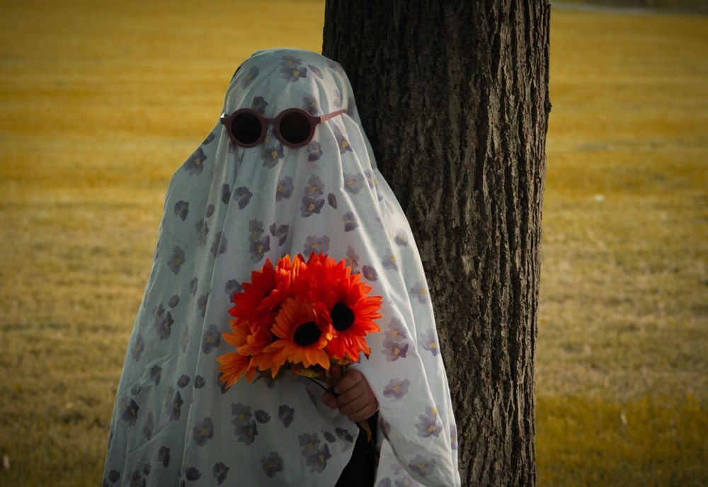 Ghost figure in floral sheet holds red flowers by tree in sunny field.