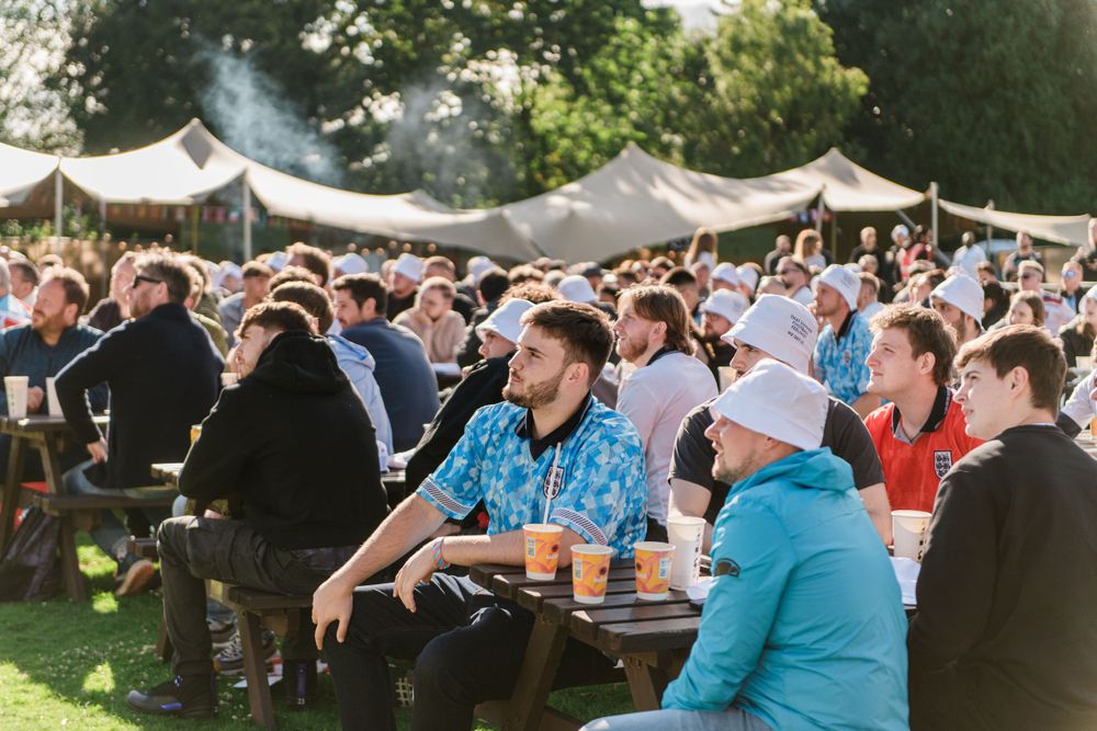 A large outdoor gathering of people seated at long tables under white canopy tents during a daytime event.