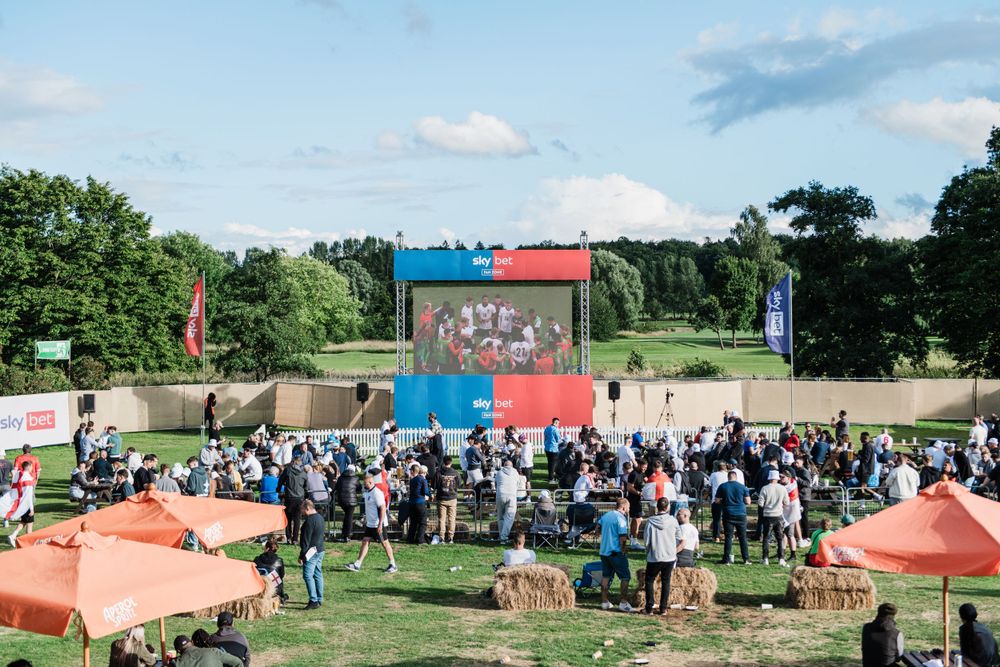 Panoramic view of an outdoor festival with colourful tents, flags, and crowds gathering on a sunny summer day.