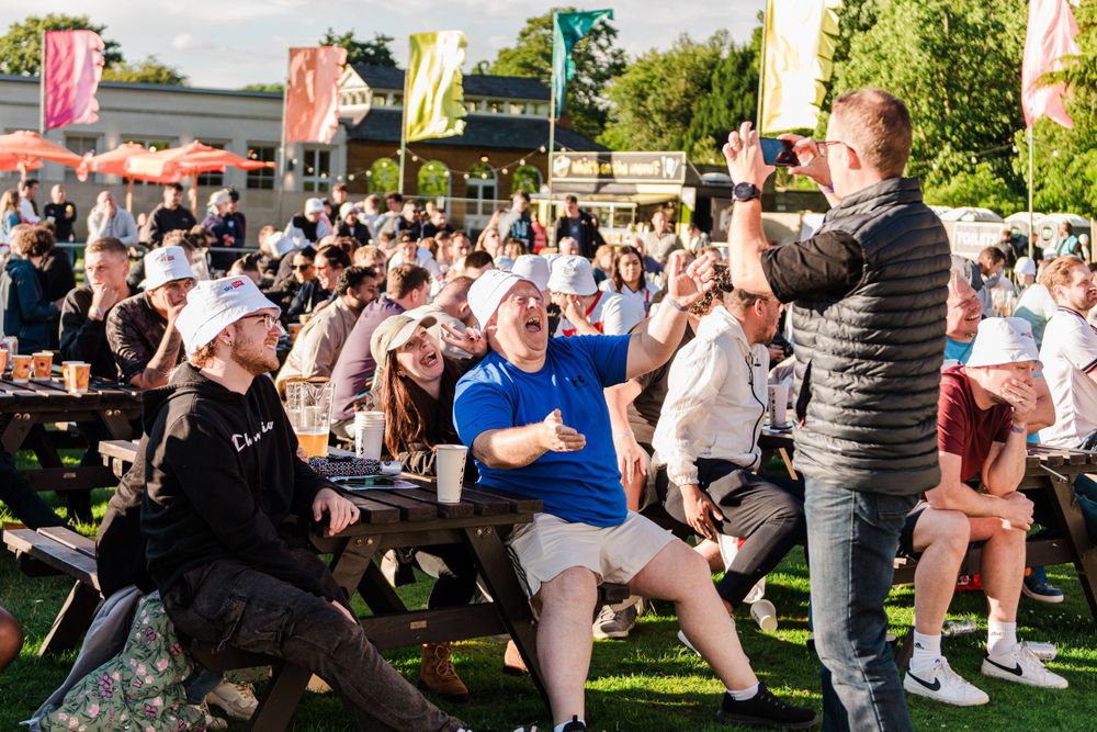 Large crowd of people dancing outdoors at a summer festival with green trees in the background.