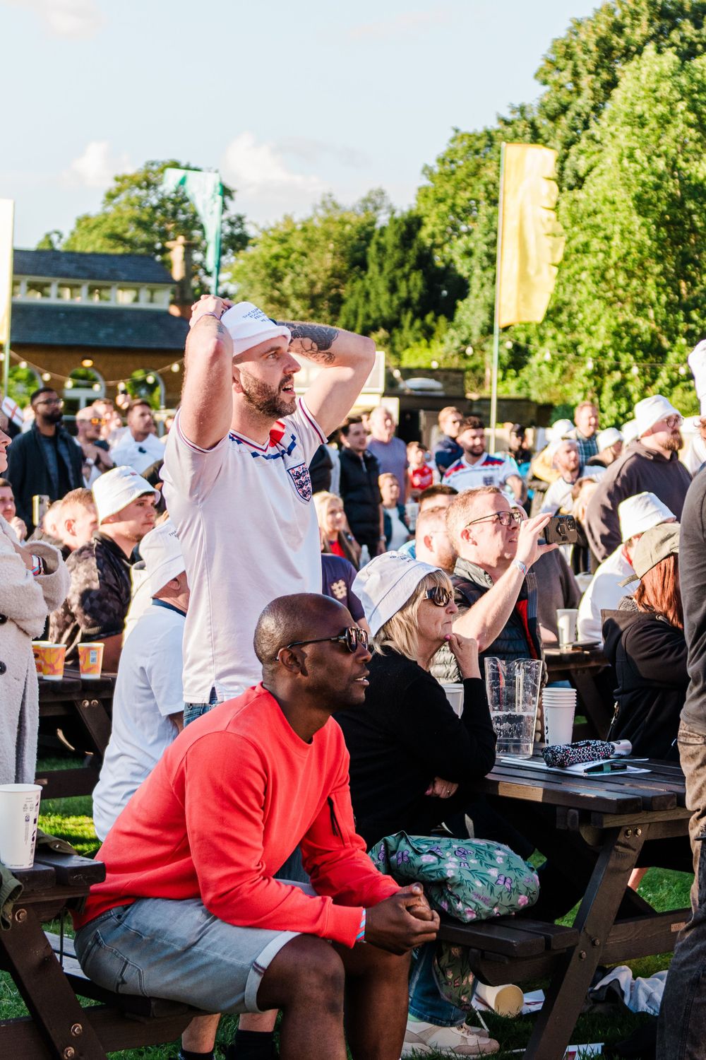 Crowd of sports fans watching and cheering at an outdoor venue with picnic tables and green trees in background.