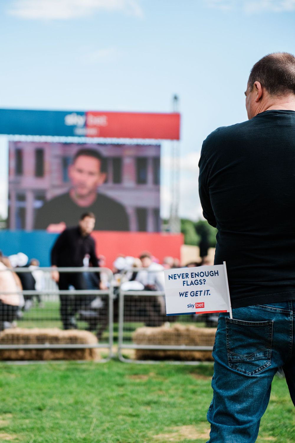 People gathered outdoors on grass for a casual event with signage and displays visible in the background.