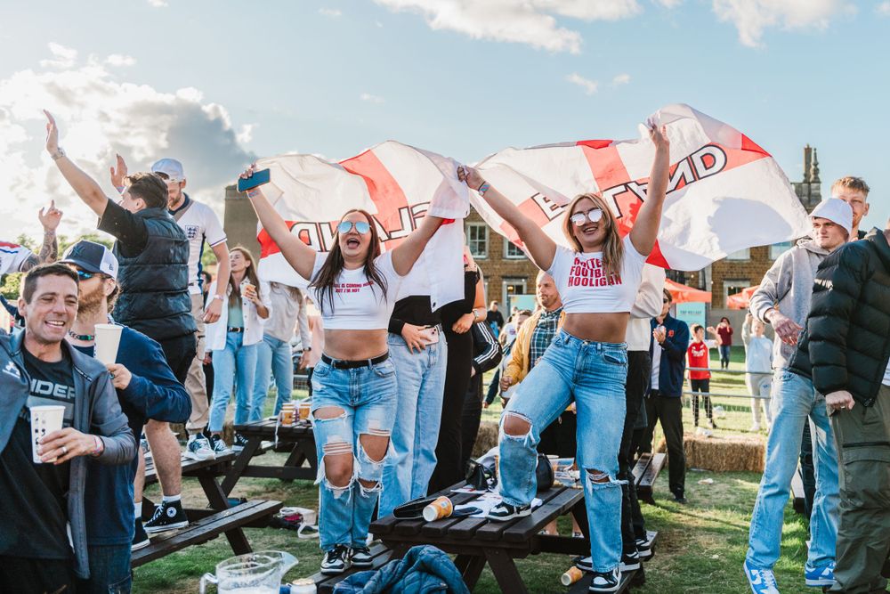 A group of students in denim jeans dance in a line formation outdoors on a sunny day with flags visible in the background.