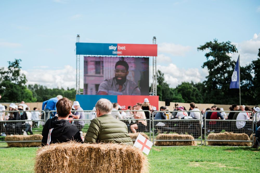 People sit on hay bales watching an outdoor event at a country fair on a sunny summer day.