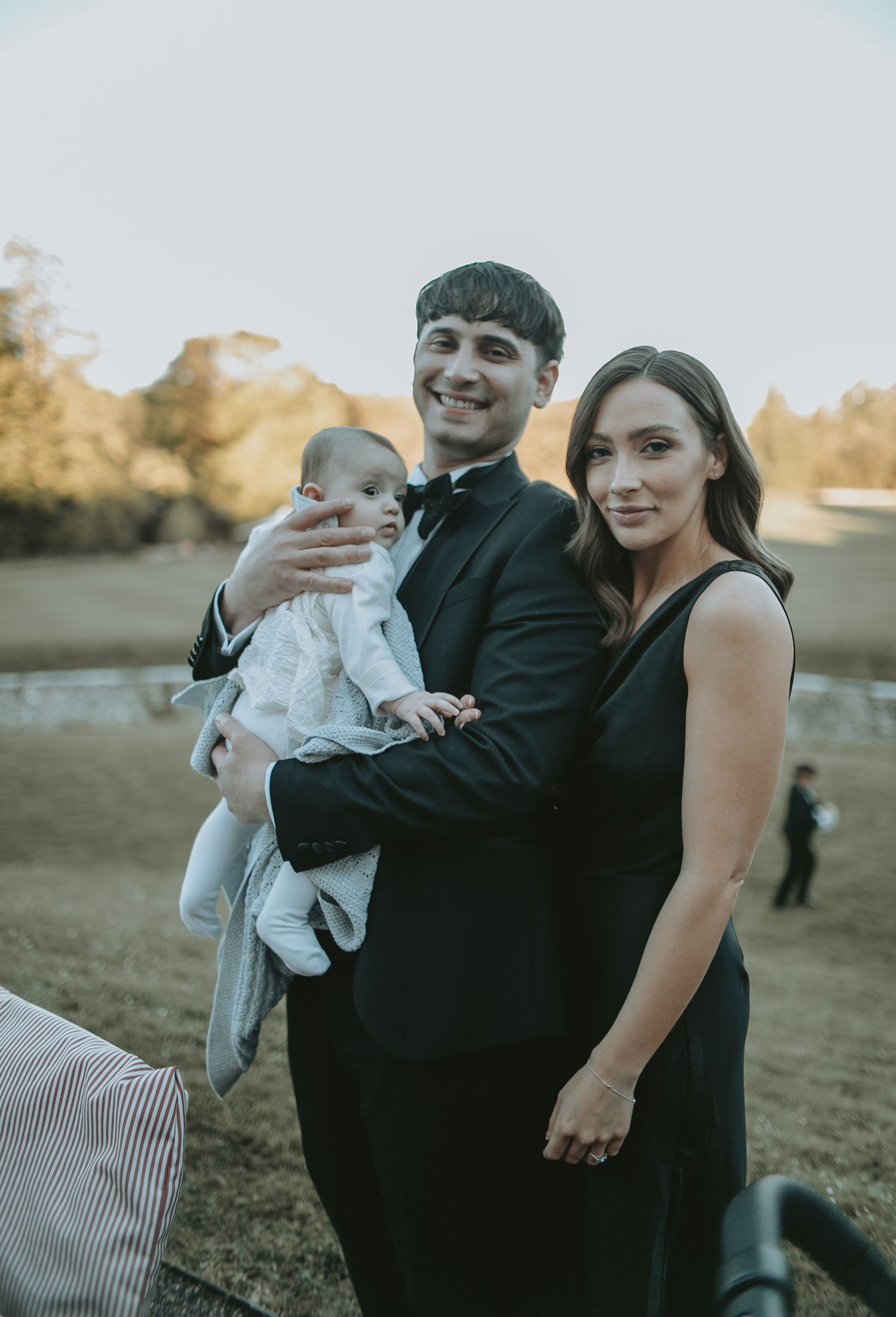 A couple in formal attire holds a baby during sunset at an outdoor wedding celebration.