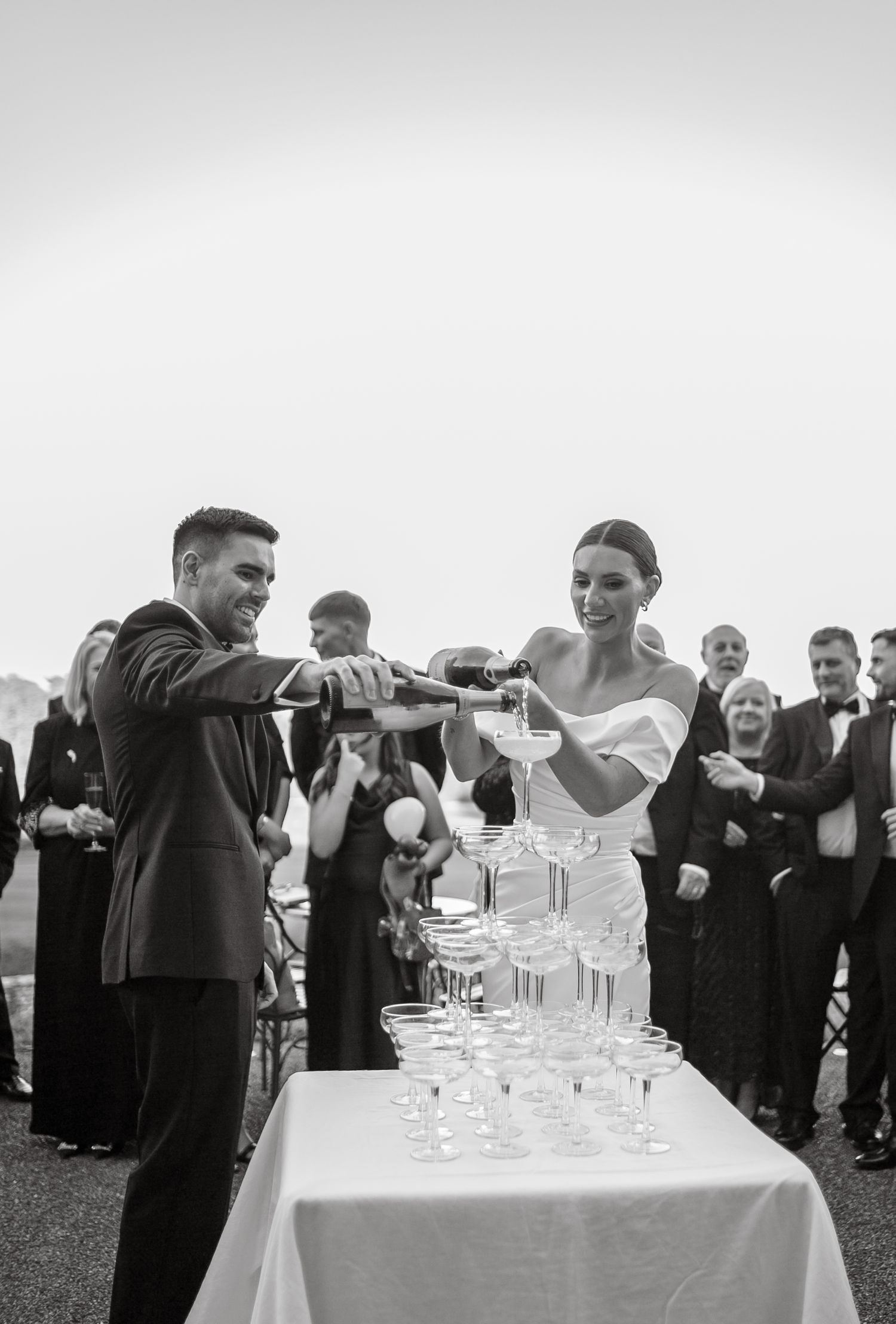 Wedding guests gather around a champagne tower at an outdoor reception.