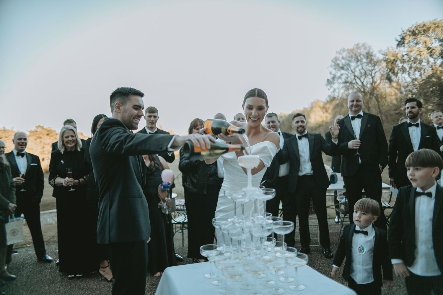 Wedding guests mingle around a champagne tower during an outdoor celebration.