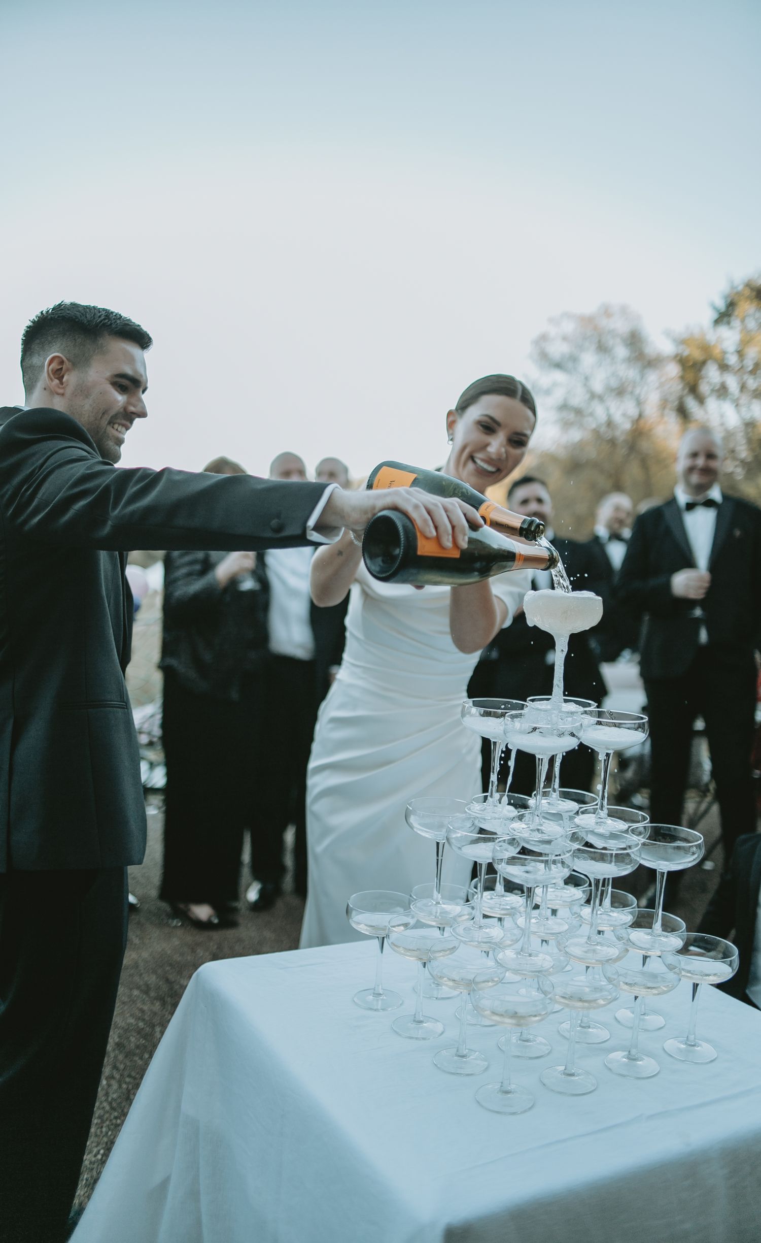 Wedding couple pouring champagne into a crystal glass tower during an outdoor celebration.