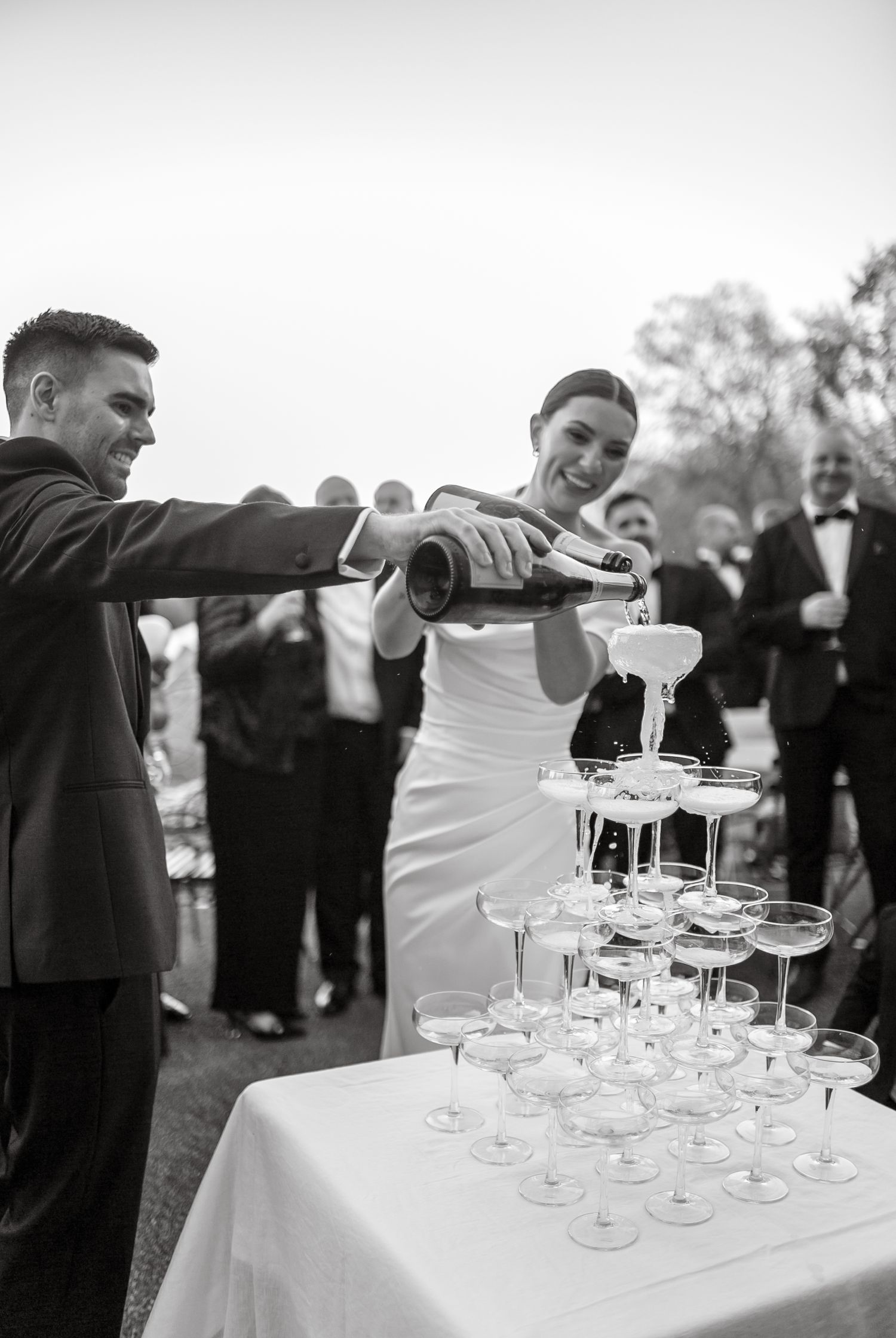 Black and white photo of newlyweds pouring champagne into a tiered glass display.