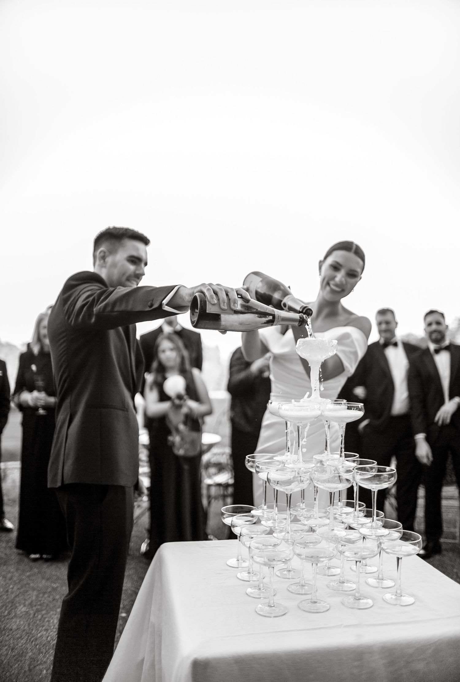 Monochrome image of a champagne tower being filled at an outdoor wedding reception.