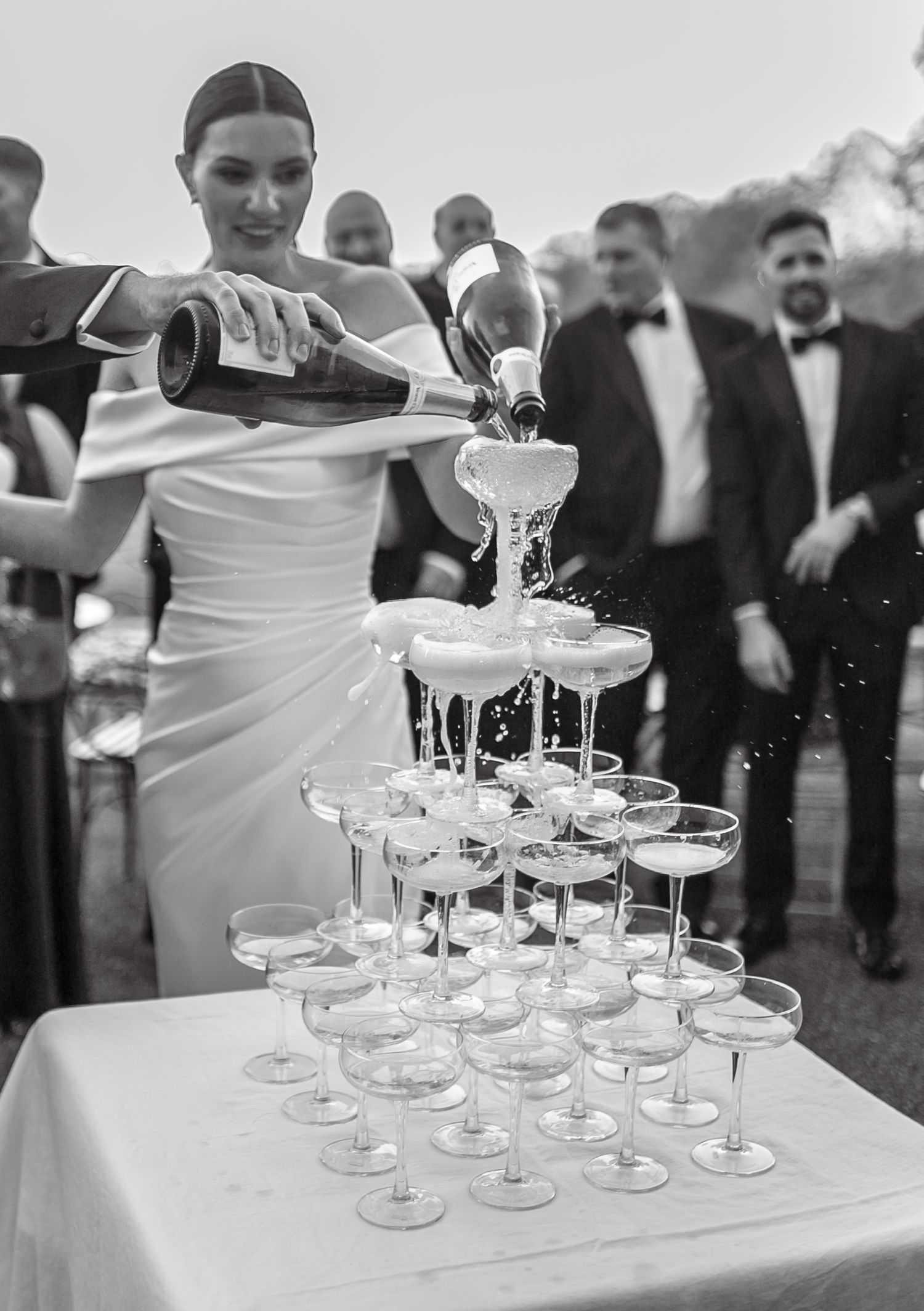 A bride in white dress pours champagne into a tower of coupe glasses at an outdoor reception.