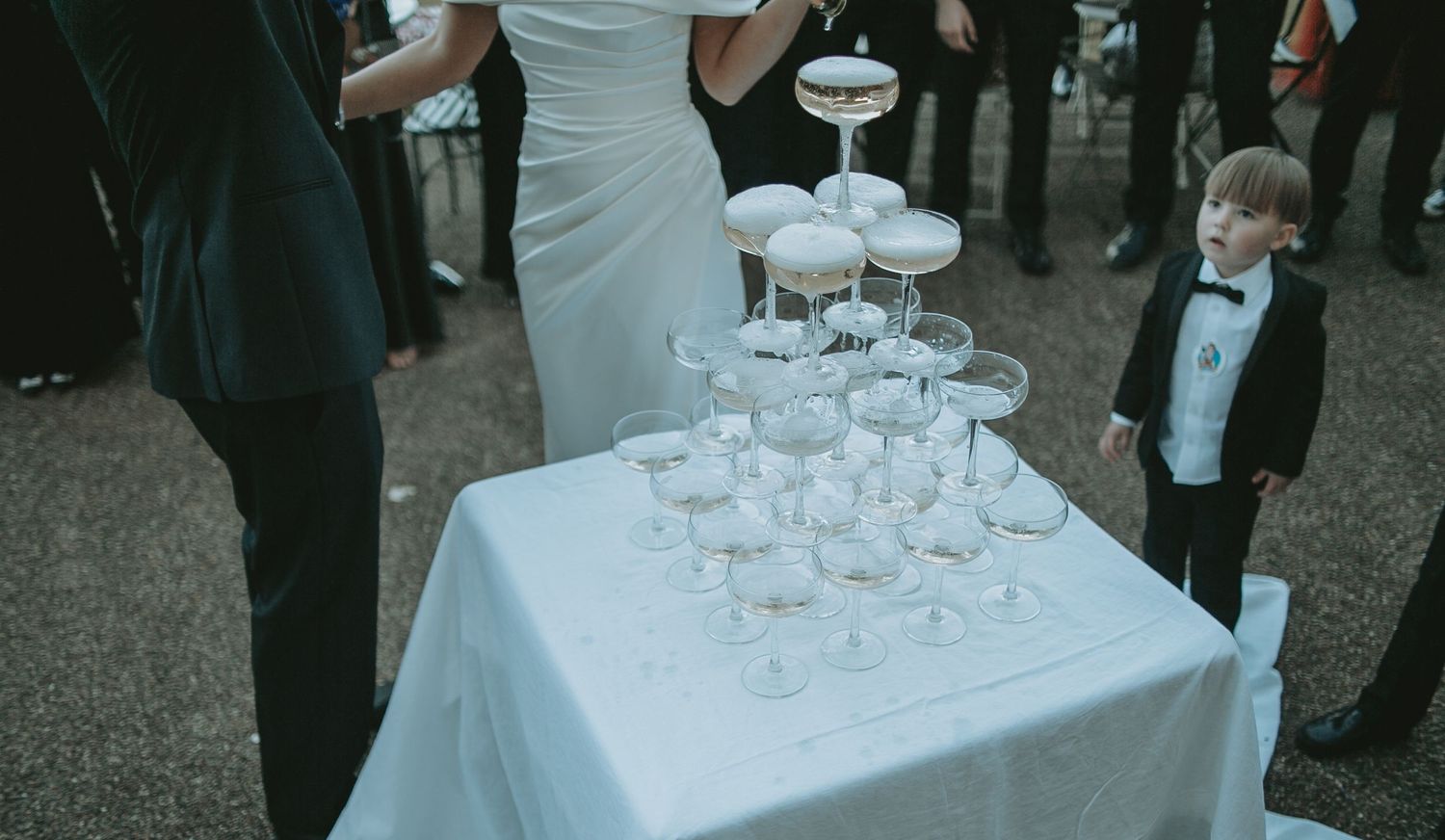 Elegant champagne tower setup on white tablecloth with wedding guests looking on.