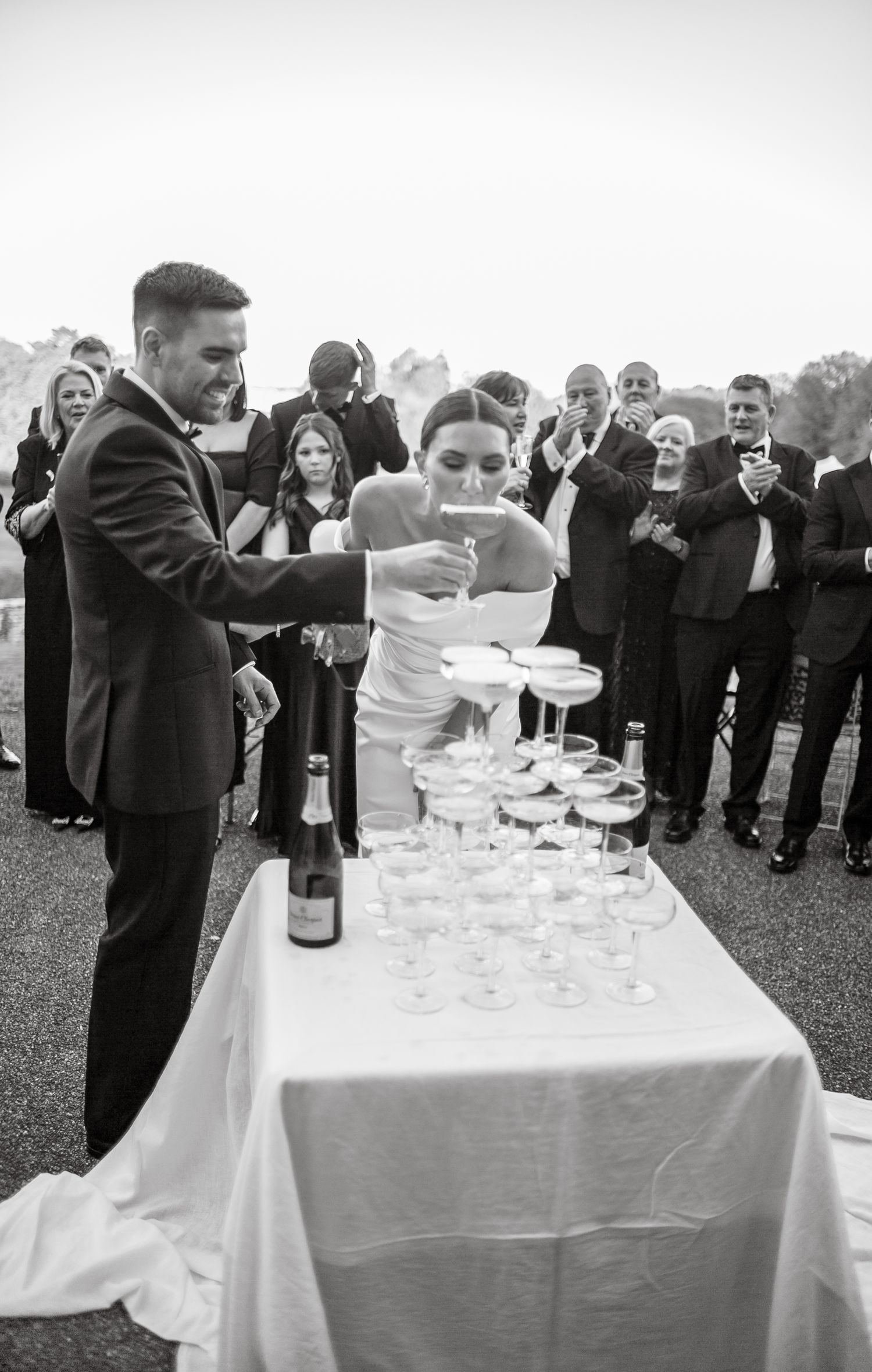 Wedding couple carefully fills a champagne tower while guests watch at outdoor reception.