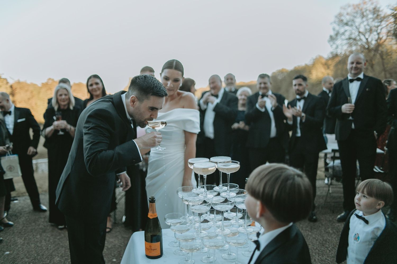 Couple pours champagne into glass tower as young ring bearers watch during outdoor celebration.