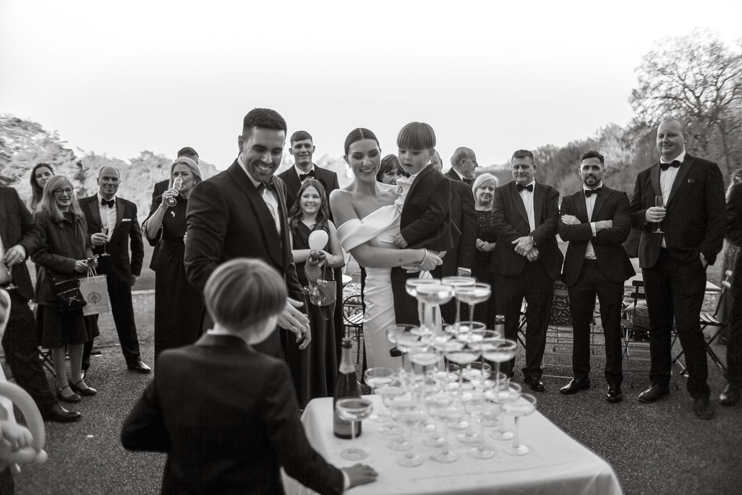 Wedding party gathers around as champagne is poured into tiered glass display outdoors.