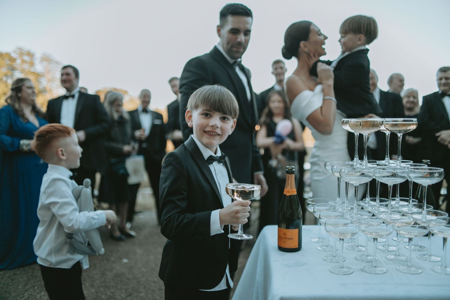 Young ring bearers in tuxedos watch as champagne tower is filled at outdoor wedding.