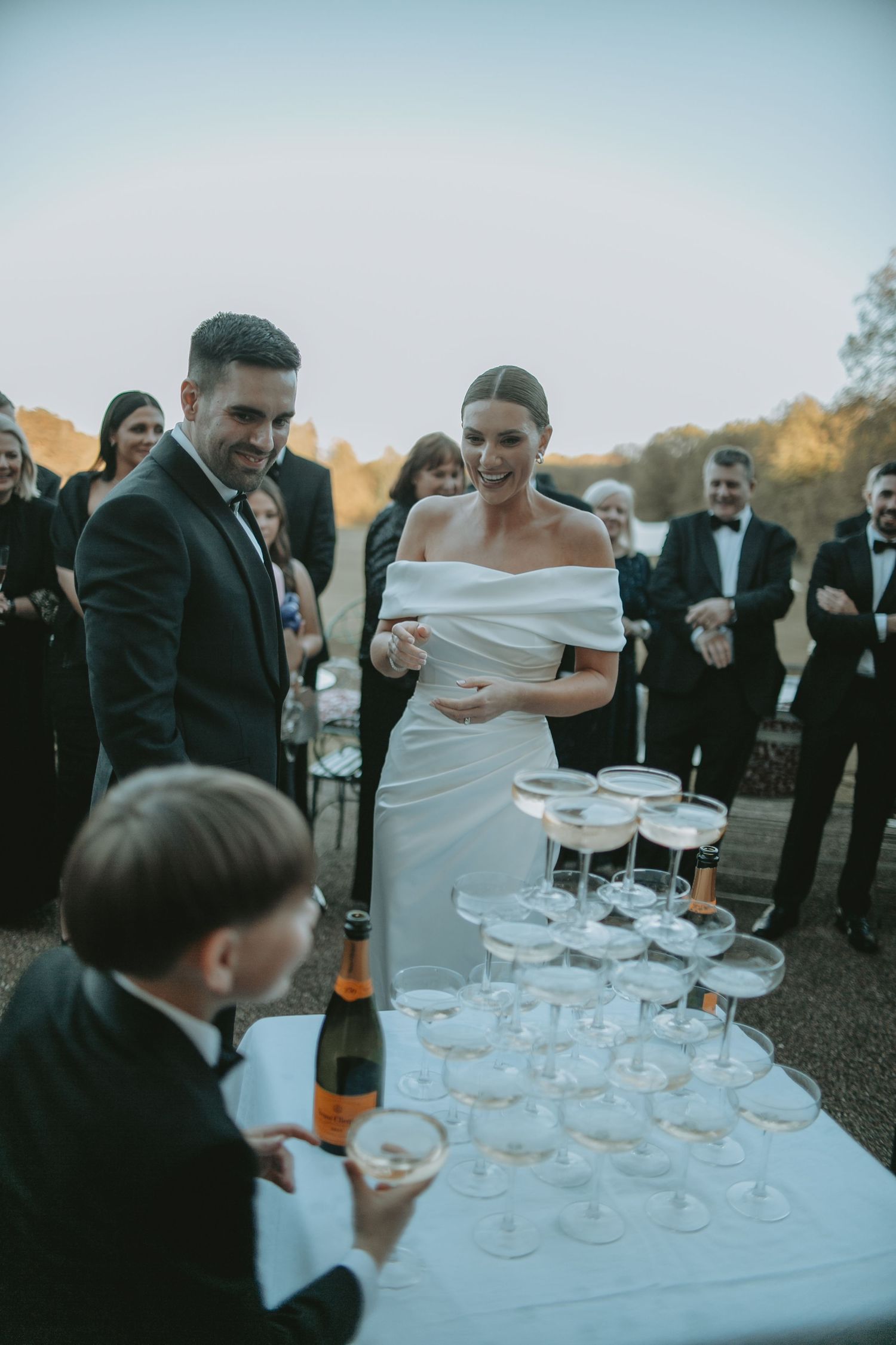 Wedding guests gather around a champagne tower being carefully poured at an outdoor reception.