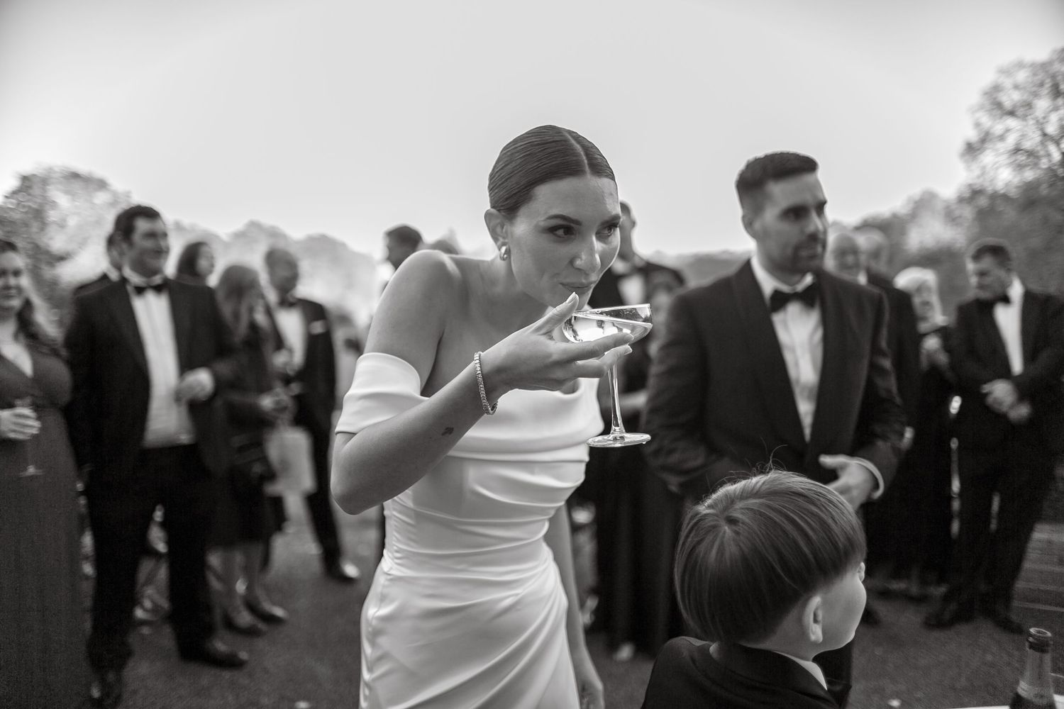 Wedding party members watch as champagne is poured into stacked crystal glasses outdoors.