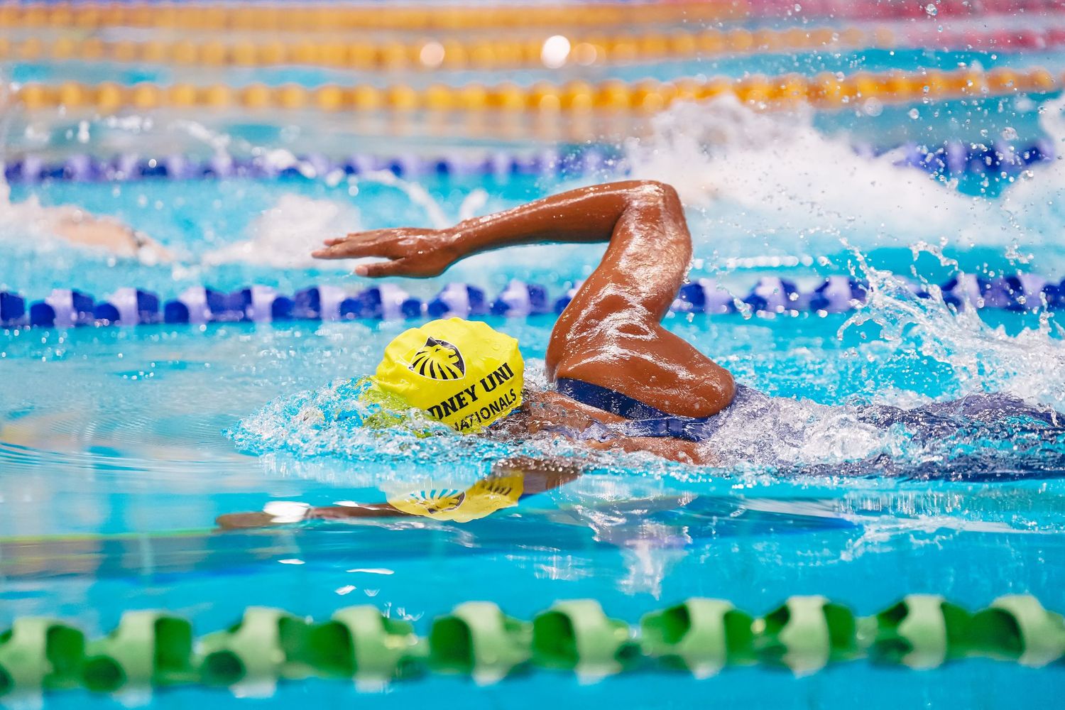 Competitive swimmer performs freestyle stroke in clear blue pool water while wearing yellow swim cap.
