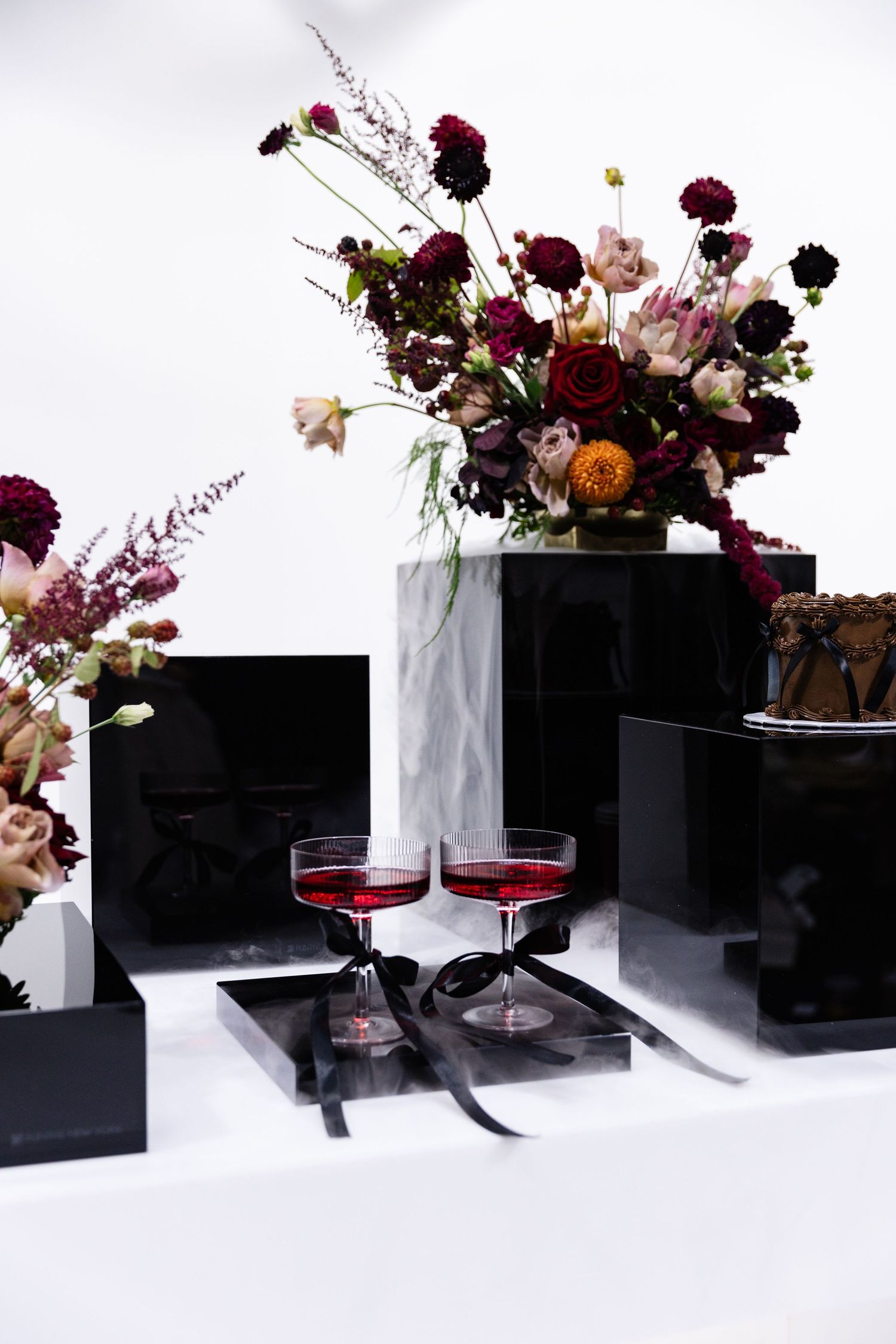 Elegant floral arrangements with deep burgundy flowers in black vases alongside wine glasses.