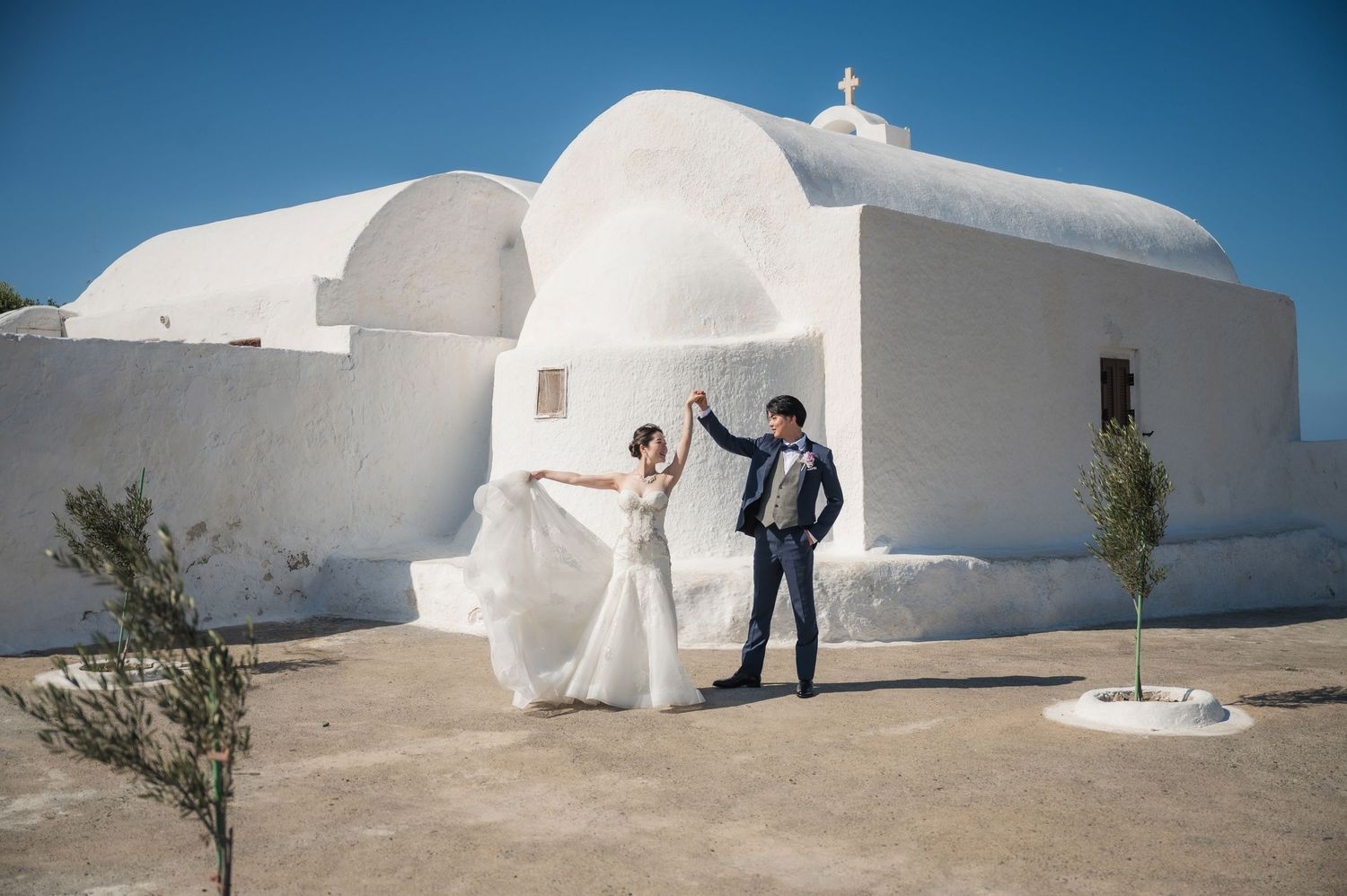 A couple poses for wedding photos in front of a traditional whitewashed Greek church with domed roof in bright sunlight.