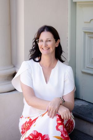 A woman in a white top and floral skirt sits smiling on outdoor steps near white architectural columns.