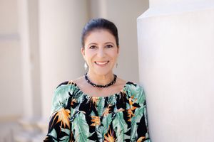 A person in a black and mint green floral print top with a choker necklace smiles against a neutral background.