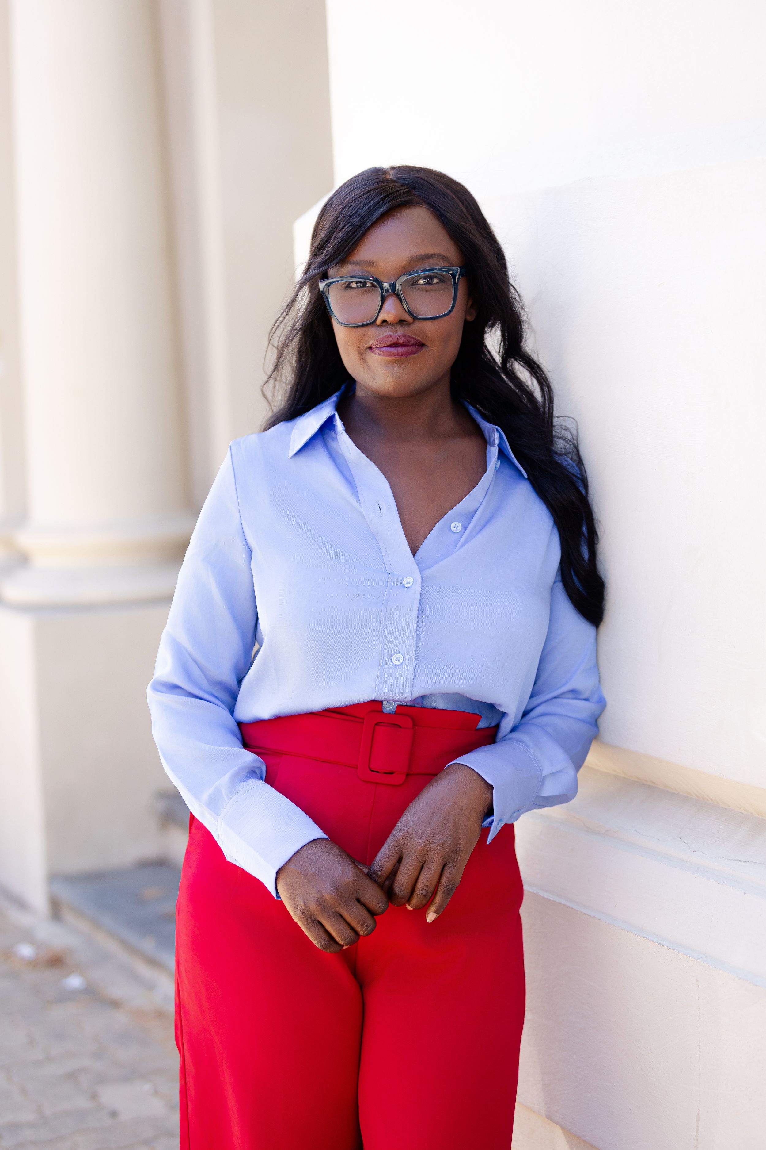 Professional portrait of someone in a light blue button-down shirt, red pants and stylish glasses standing against a pillar.