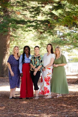 A group of women in colorful dresses standing together under tall evergreen trees in a park setting.