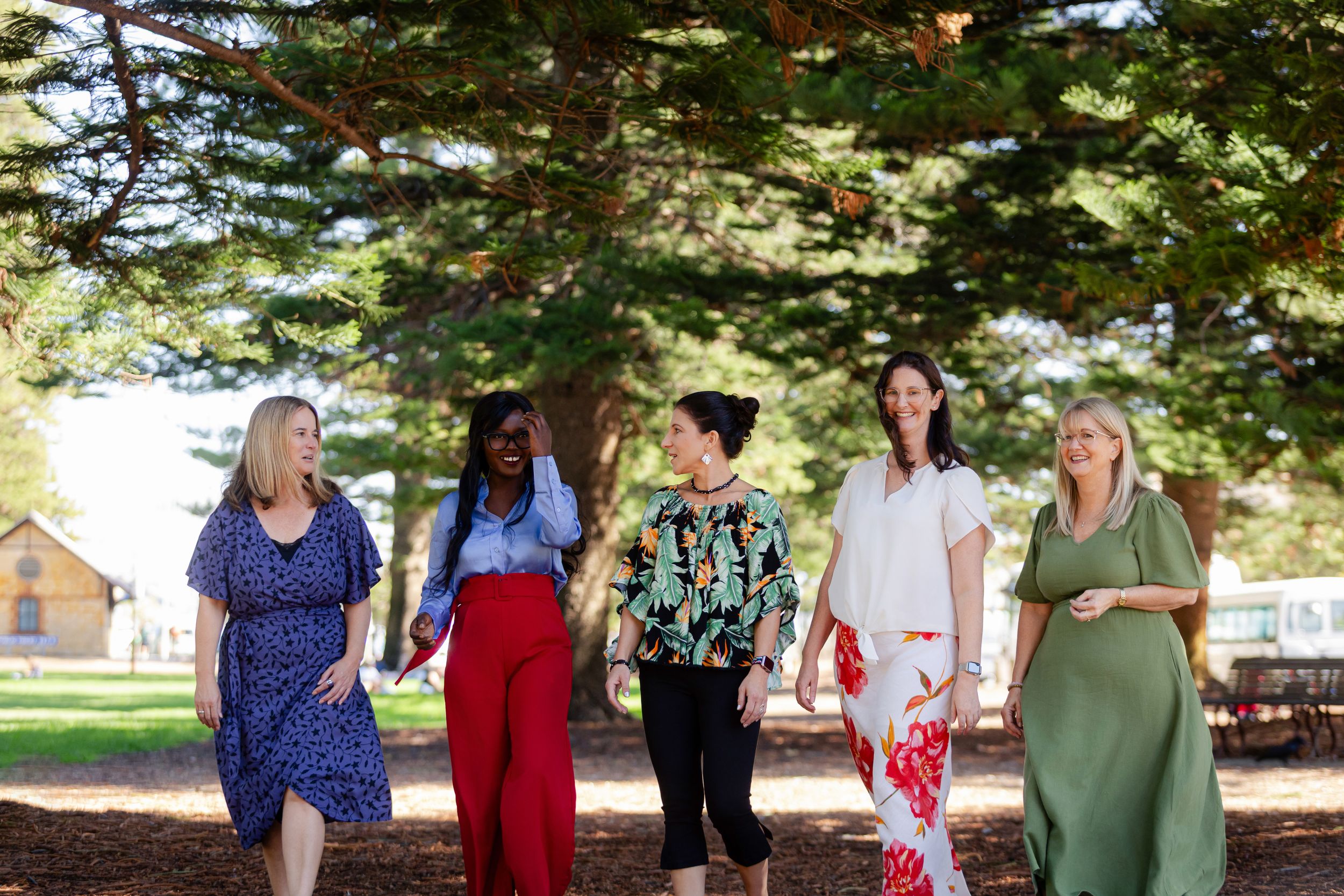 Five friends walk together outdoors enjoying a beautiful sunny day under tree canopy at a park.
